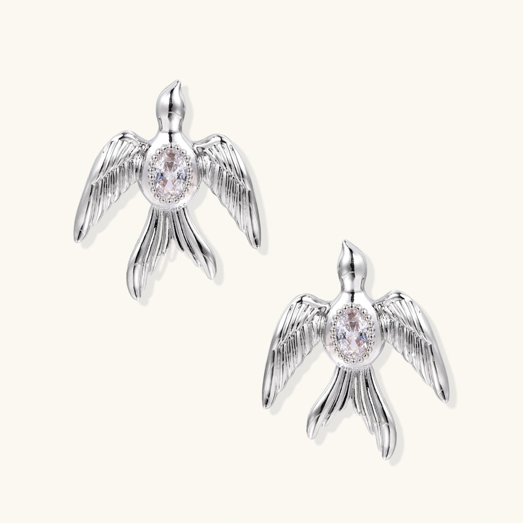 White gold-plated bird earrings with spread wings and oval zirconia center on cream background