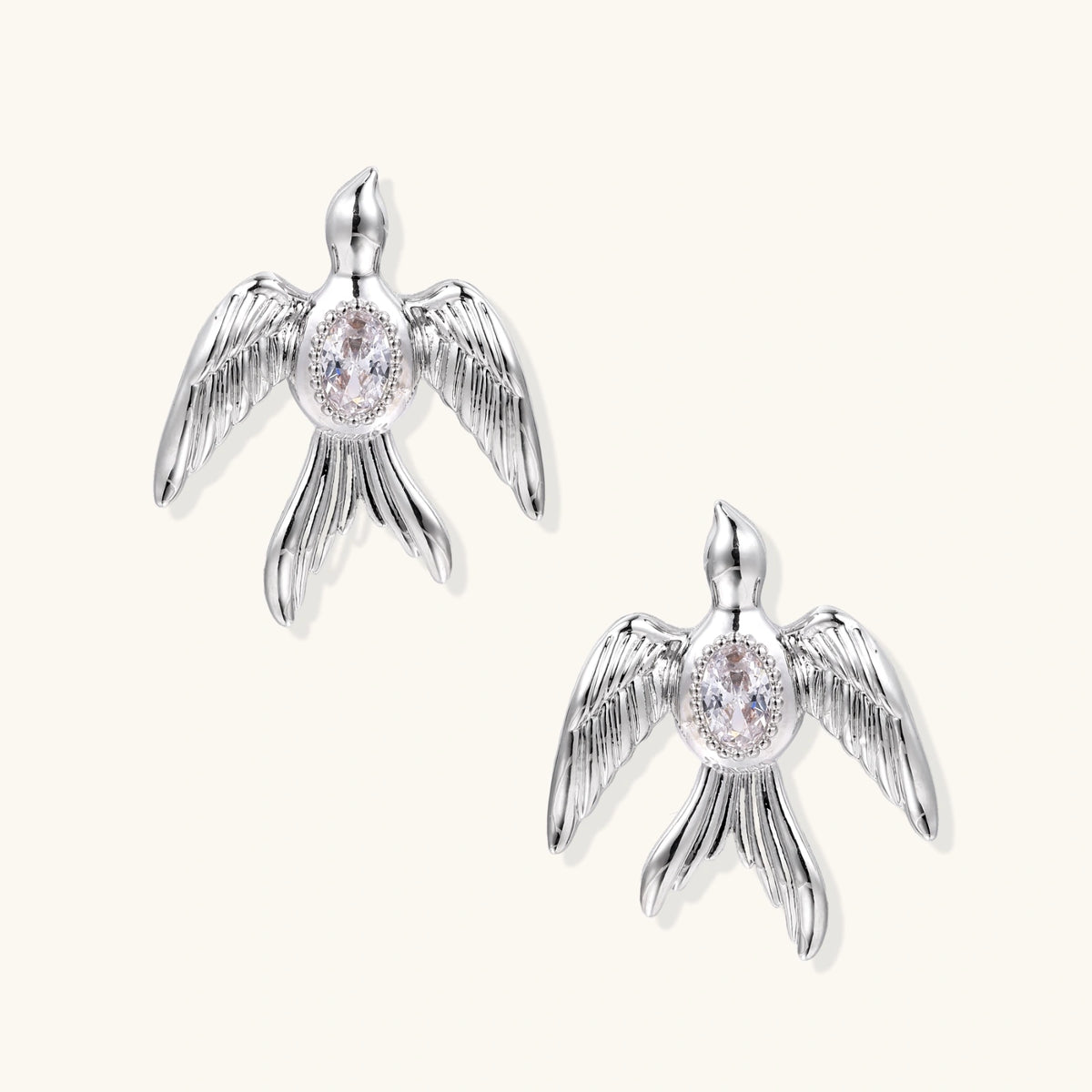 White gold-plated bird earrings with spread wings and oval zirconia center on cream background