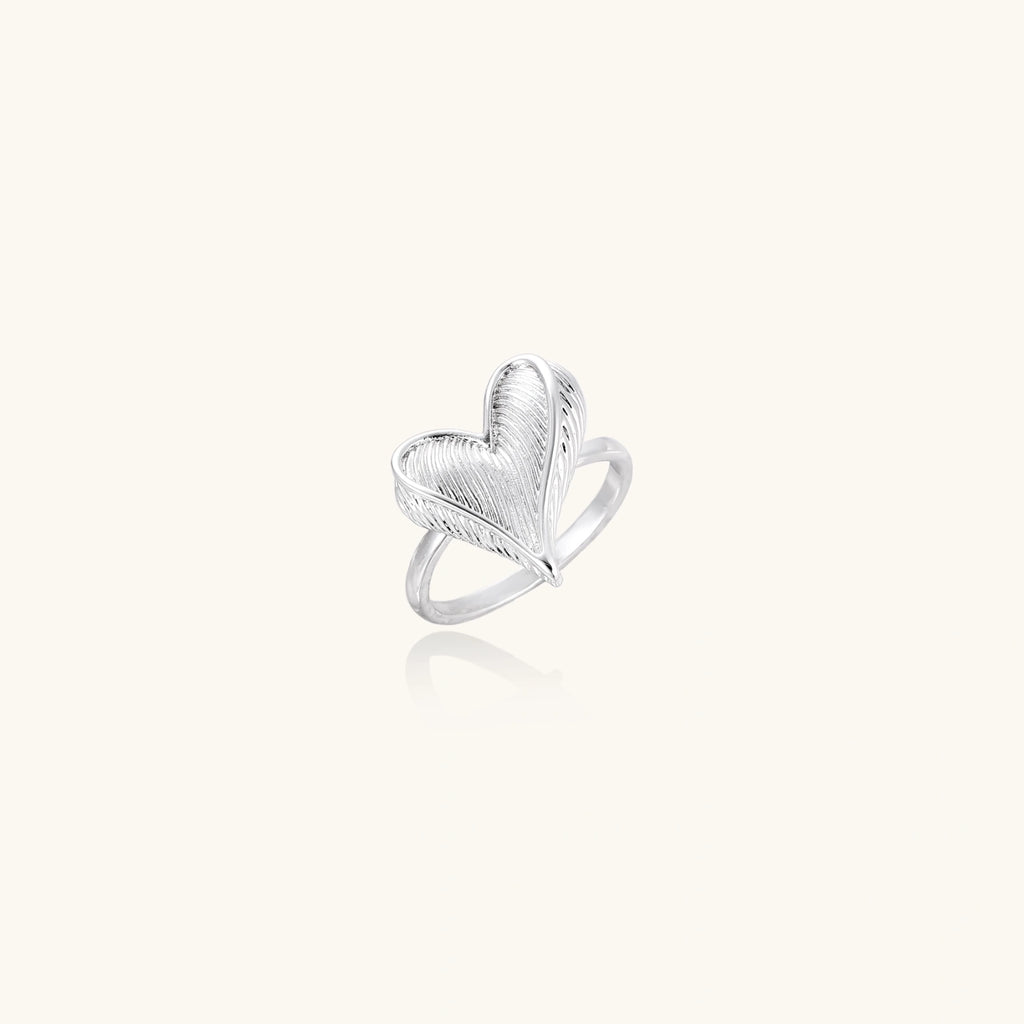 Silver ring with 3D heart formed by textured feather-like elements, white gold plated eco-brass on neutral background.